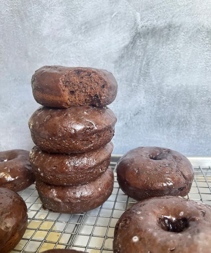 A stack of chocolate donuts with a few more on the side on a wire rack.