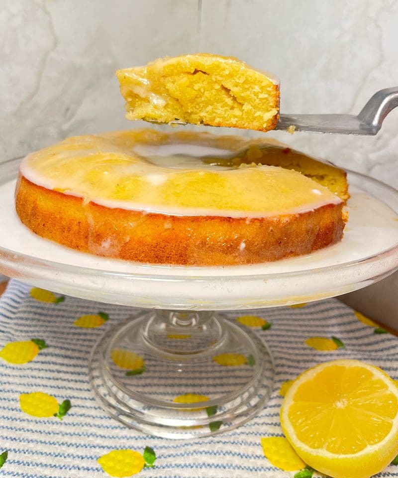 A slice of lemon cake being lifted from a whole cake on a glass stand.